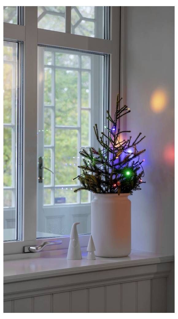 Kleiner Weihnachtsbaum mit bunten Lichtern in einem weißen Topf auf der Fensterbank. Das Fenster zeigt einen Blick auf Bäume draußen.