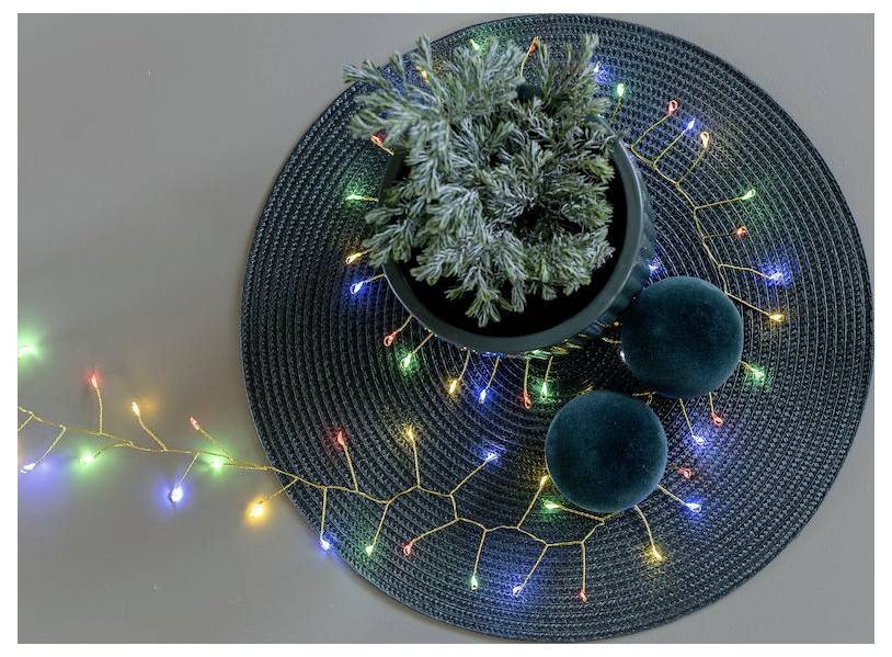 Ein kleiner Topf-Nadelbaum steht auf einer gewebten Matte mit bunten Lichterketten und zwei dunkelgrünen runden Kissen, die eine festliche Dekoration schaffen.