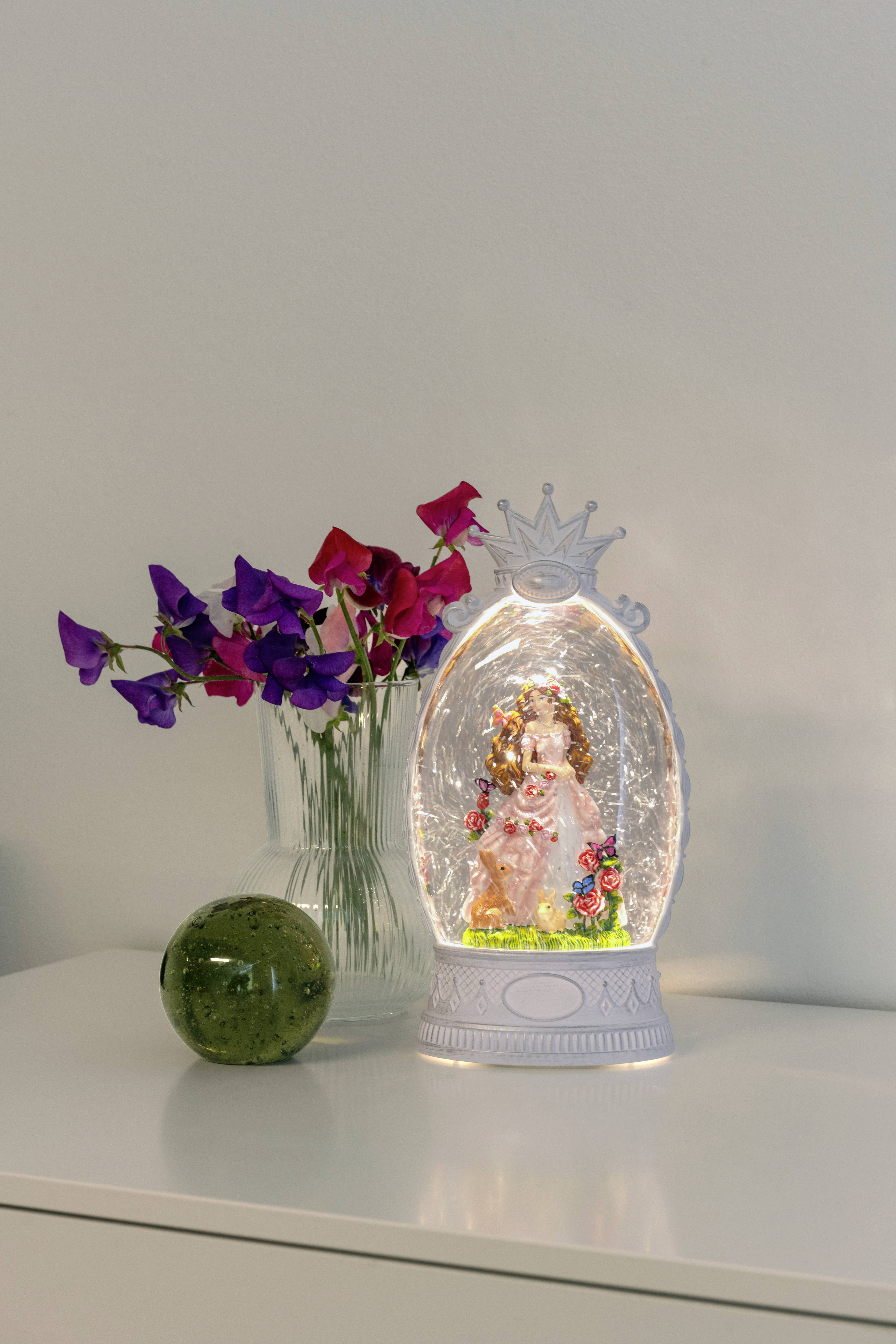 Eine dekorative Leuchte mit einer Engelsfigur im Inneren, neben einer Vase mit rosa und lila Blumen und einer kleinen grünen Glaskugel auf einer weißen Oberfläche.