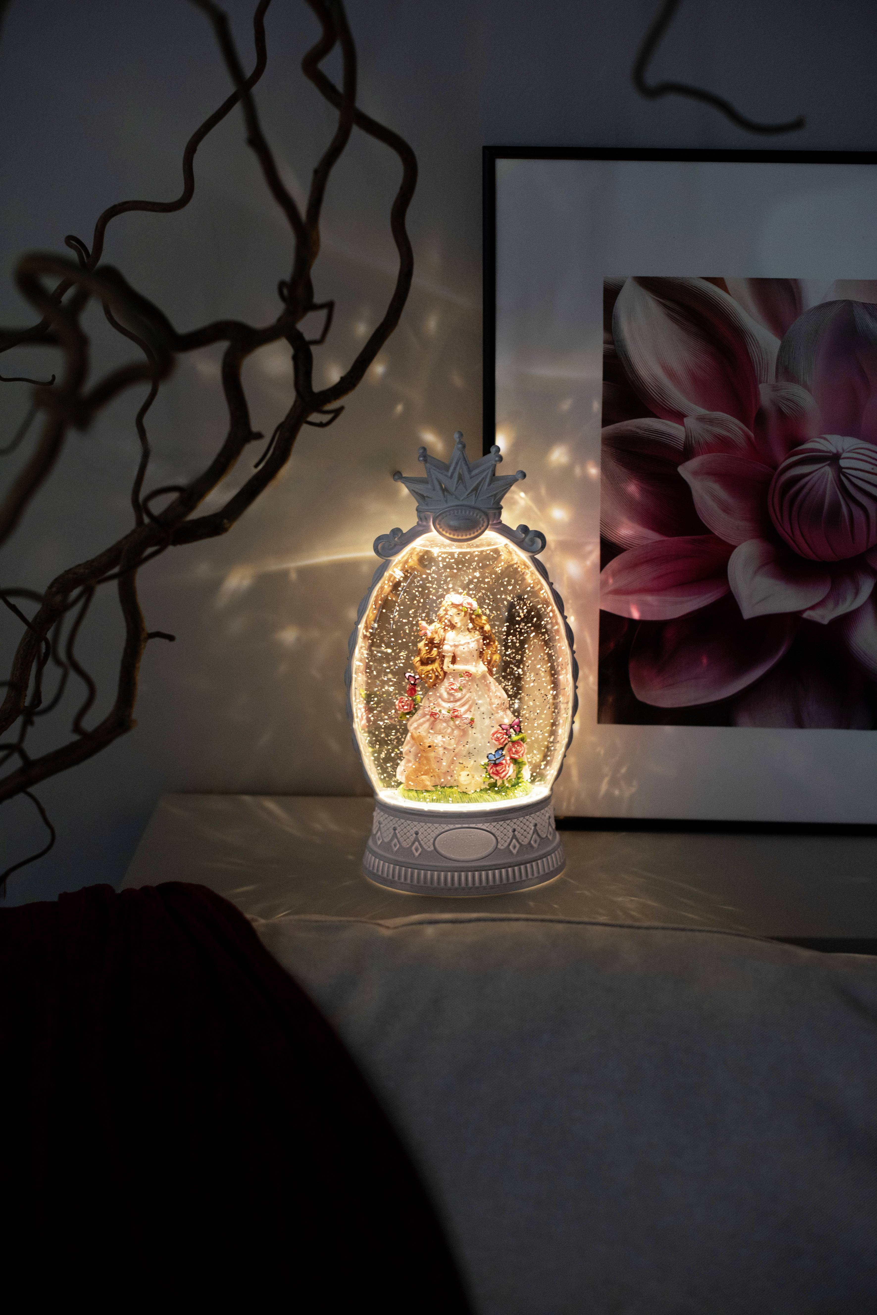 Eine leuchtende Schneekugel steht auf einem Tisch und zeigt eine Prinzessinfigur im Inneren. Daneben hängt an der Wand ein Kunstwerk mit einer rosa Blume.