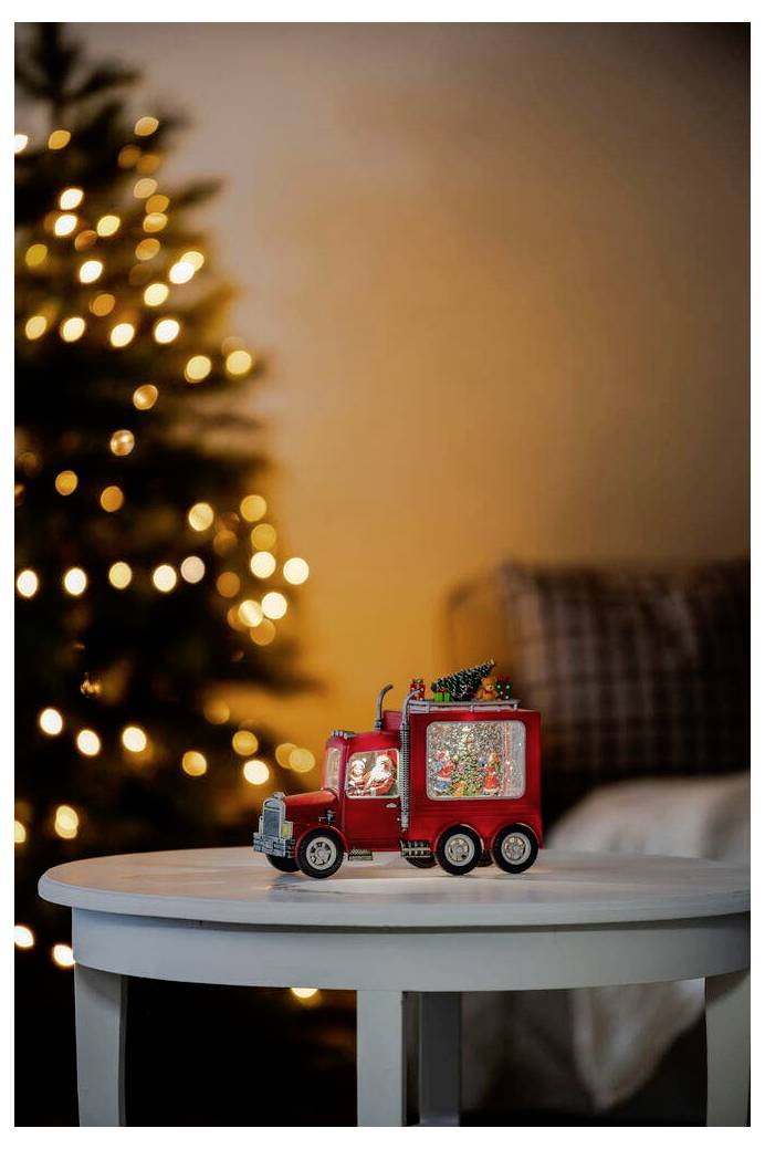 Ein kleiner roter Spielzeug-Lastwagen mit Weihnachtsbeleuchtung steht auf einem weißen Tisch, während ein unscharfer, beleuchteter Weihnachtsbaum im Hintergrund eine festliche Szenerie schafft.