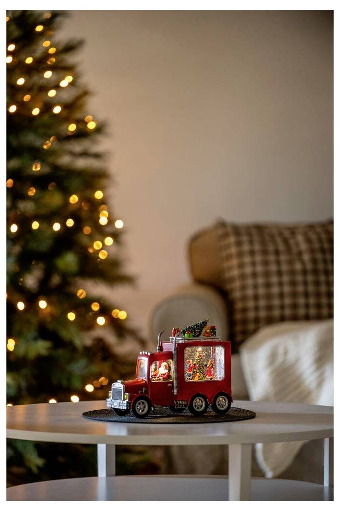 Ein roter Spielzeug-Lastwagen mit festlicher Dekoration steht auf einem Tisch vor einem beleuchteten Weihnachtsbaum und verbreitet eine gemütliche Feiertagsstimmung im Wohnzimmer.