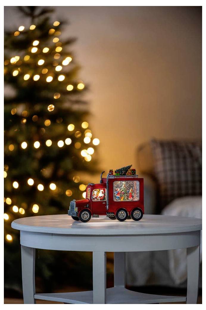 Ein roter Spielzeug-Lastwagen mit Weihnachtsdekoration steht auf einem runden weißen Tisch. Ein mit Lichtern geschmückter Weihnachtsbaum befindet sich im Hintergrund.