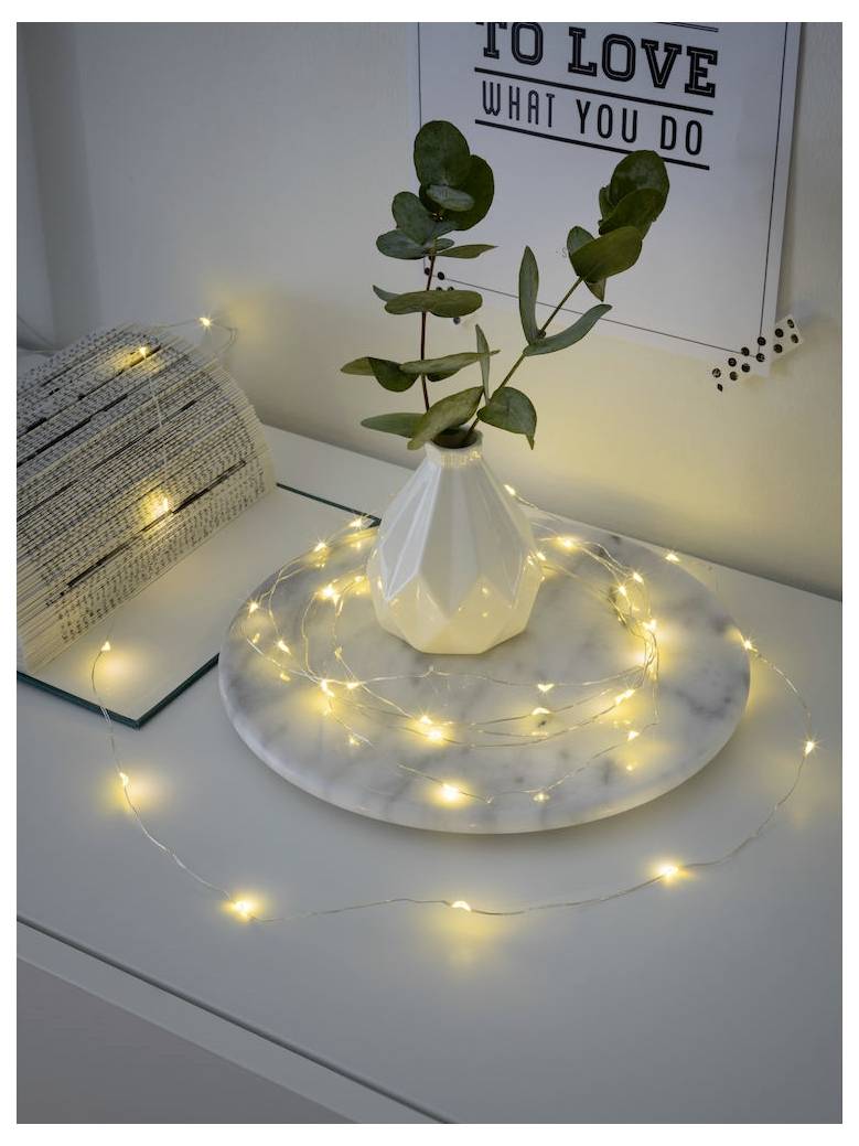 Eine weiße Vase mit grünen Blättern und ein Marmortablett, geschmückt mit Lichterketten auf einem Tisch. Ein Schild im Hintergrund trägt die Aufschrift ‚LIEBE DAS, WAS DU TUST'.