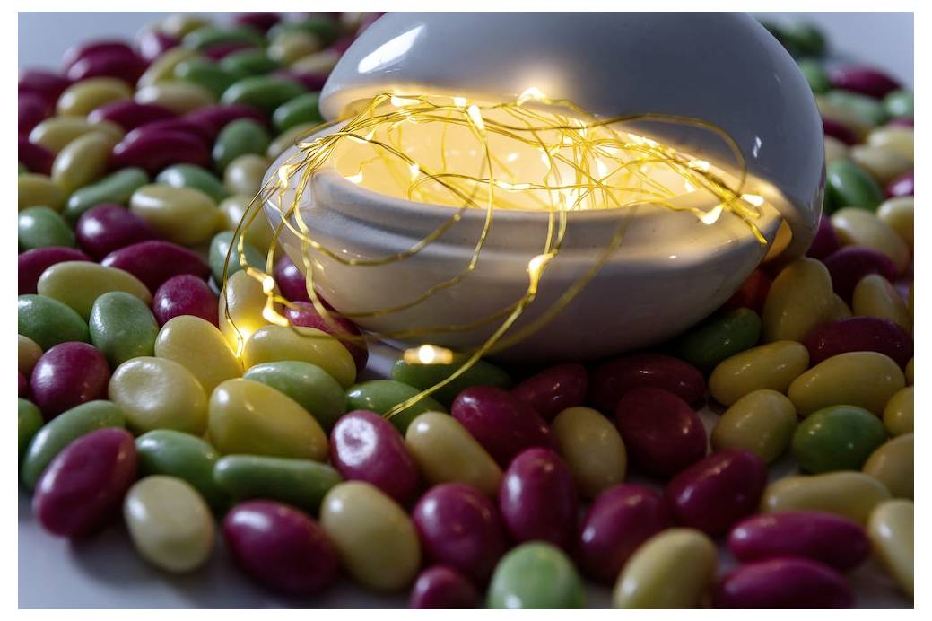 Eine weiße Keramikkugel, teilweise geöffnet, mit leuchtenden Feenlichtern im Inneren, umgeben von bunten Gummibärchen.