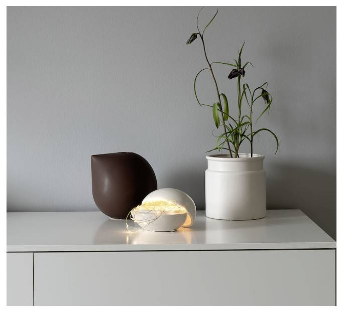 Eine minimalistische Regaldekoration mit einer kleinen leuchtenden Lichtskulptur, einer braunen Vase und einer weißen Vase mit einer blattreichen Pflanze vor einer grauen Wand.