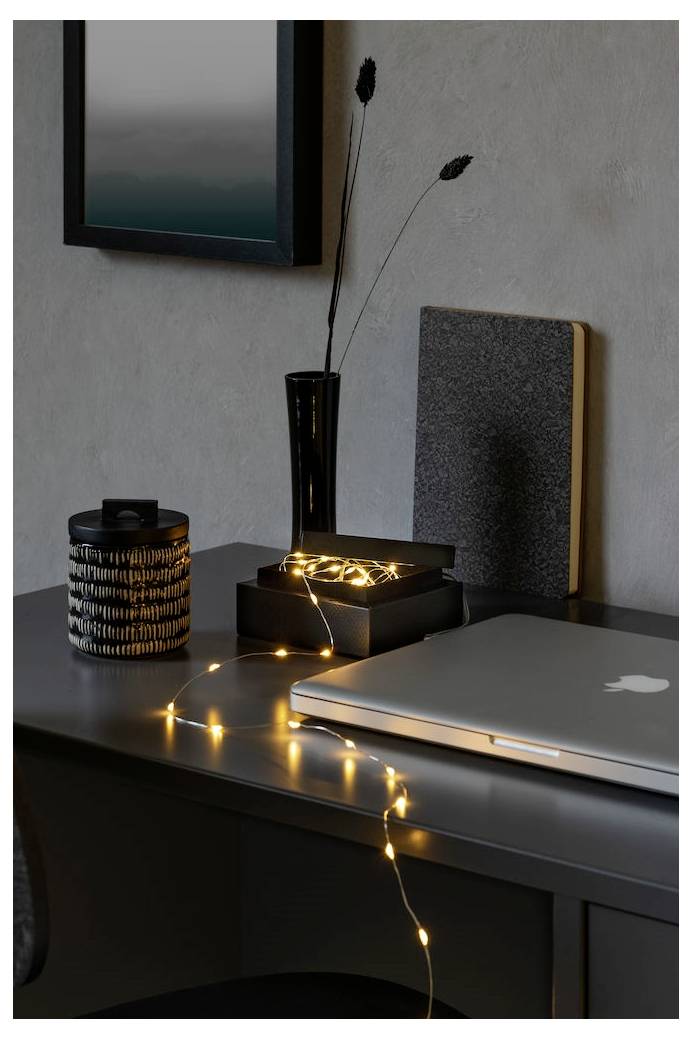 Ein Laptop steht auf einem dunklen Schreibtisch mit warmen Lichterketten, einer schwarzen Vase mit dünnen Zweigen und einigen dekorativen Gegenständen in einem minimalistischen Ambiente.