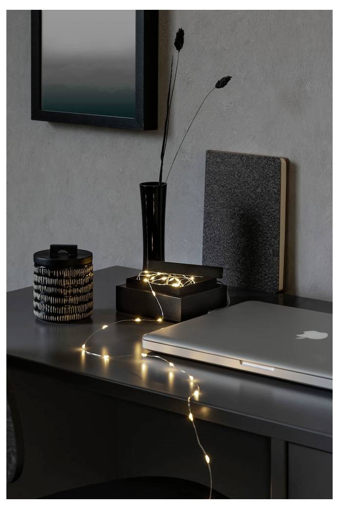 Ein Schreibtisch mit einem geschlossenen Laptop, einem Behälter, Lichterketten, einer schwarzen Vase mit getrockneten Blumen und einem gerahmten Bild an der Wand.