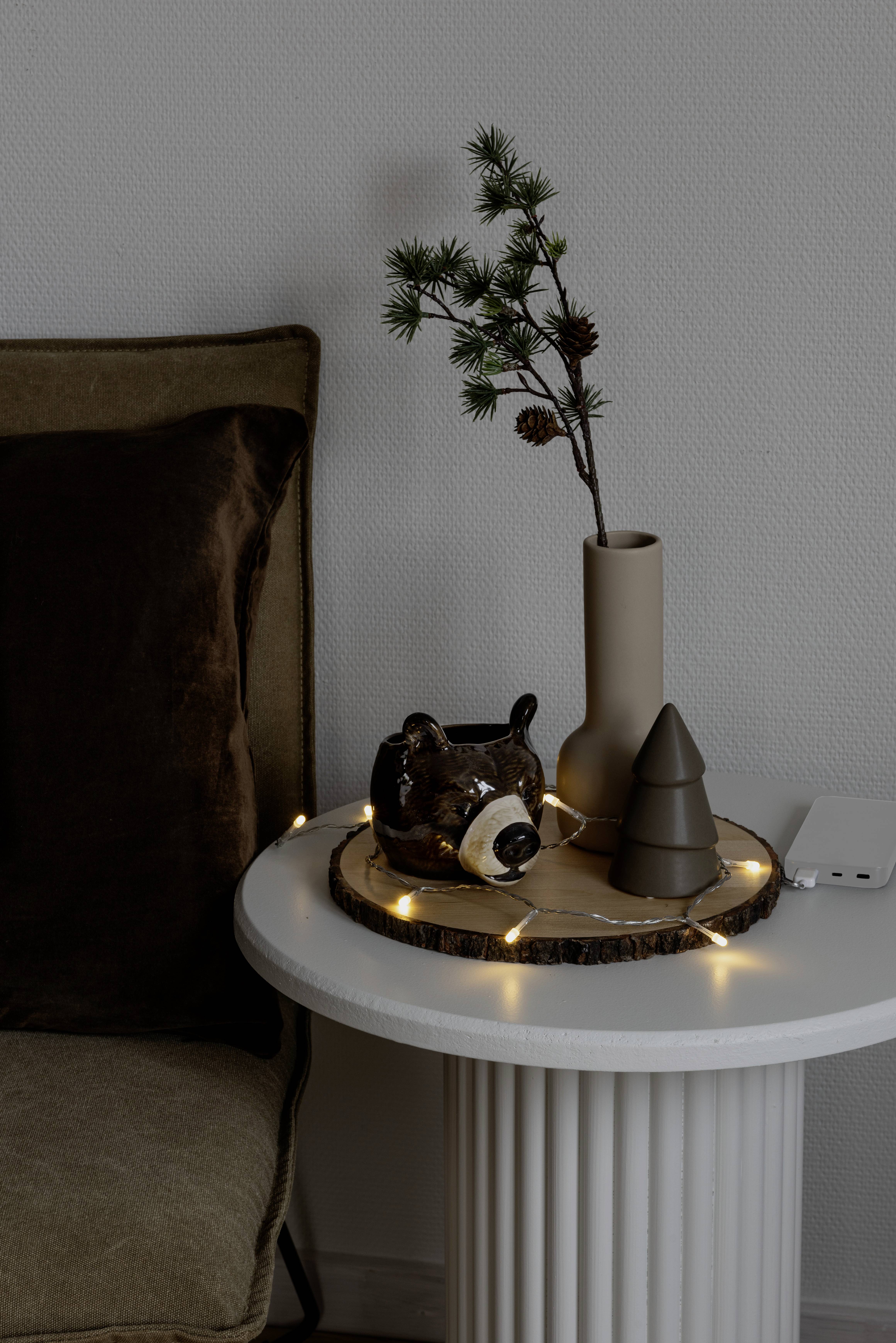 Ein kleiner Tisch trägt einen dekorativen Bärenkrug, eine Vase mit Zweigen, eine baumförmige Dekoration und Lichterketten und schafft dadurch eine gemütliche Atmosphäre.