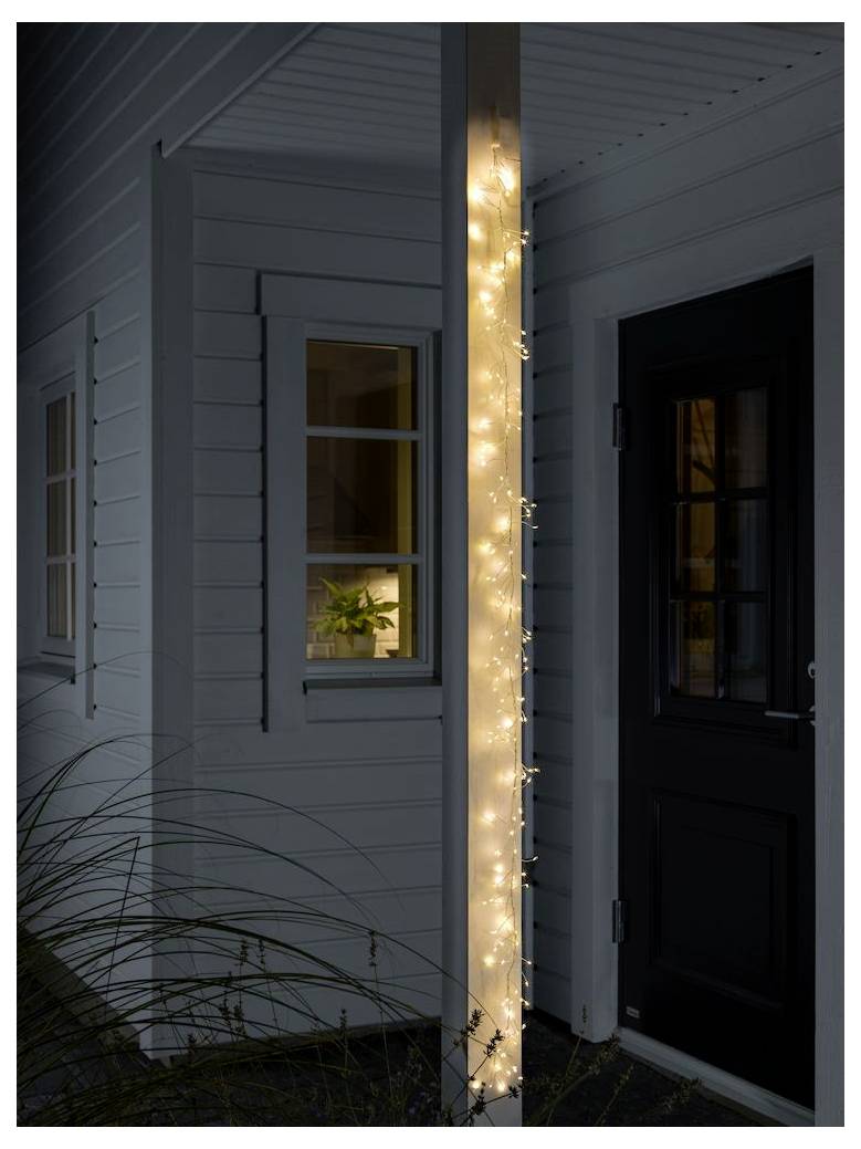 Eine Säule auf der Veranda eines Hauses ist in leuchtende Lichterketten gehüllt. Daneben lässt ein warmbeleuchtetes Fenster eine Topfpflanze im Inneren erkennen.