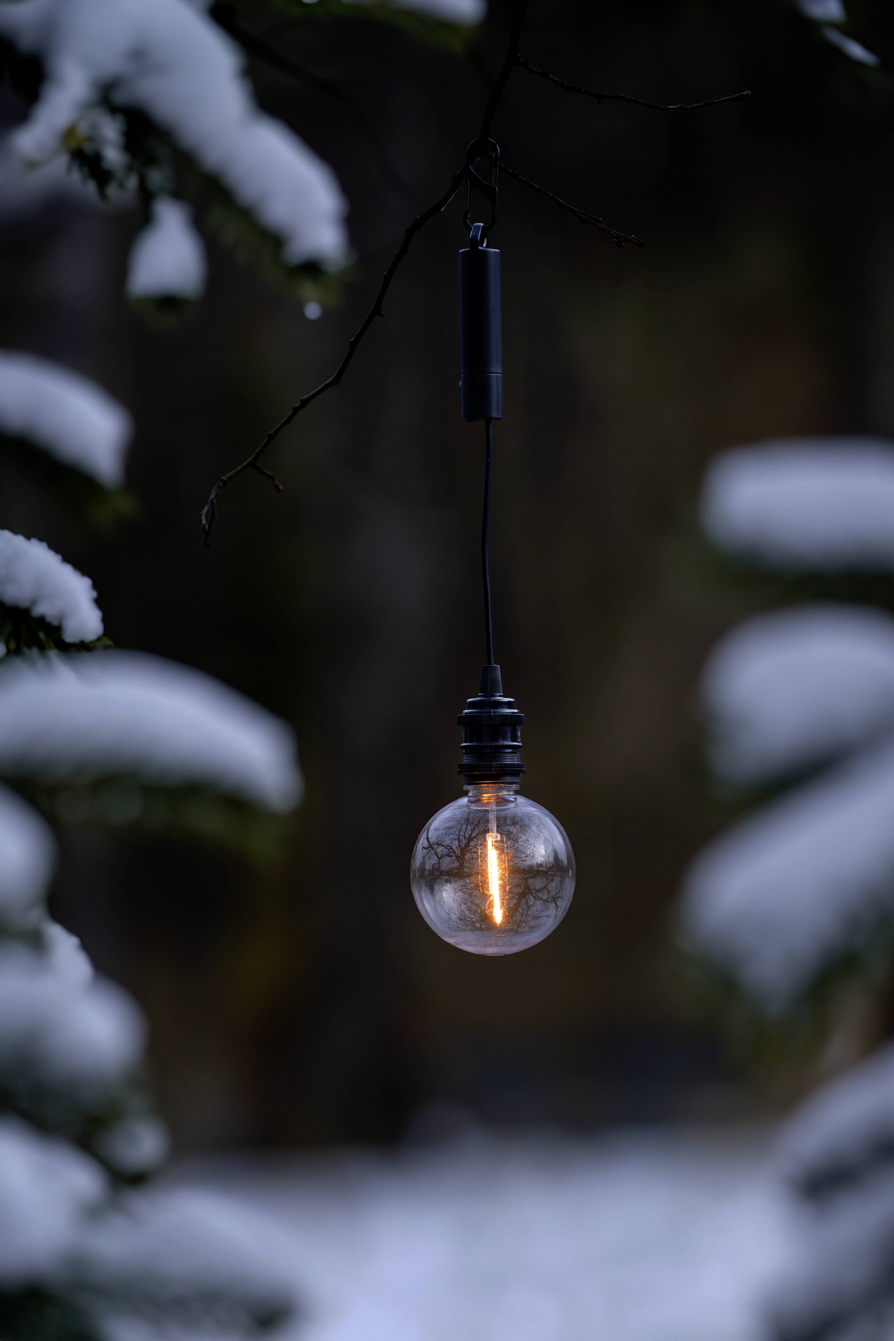 Eine leuchtende Glühbirne hängt an einem Draht in einer verschneiten Außenumgebung, mit verschwommenen schneebedeckten Ästen im Hintergrund.