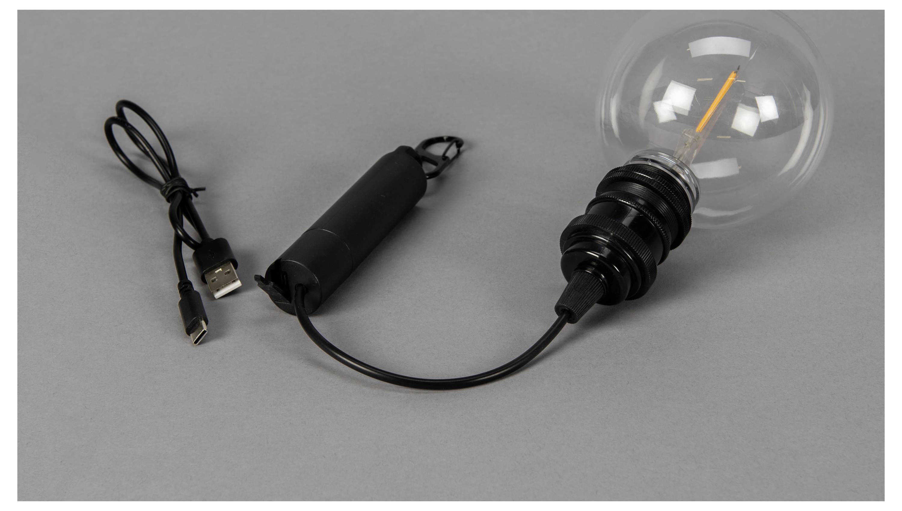 Eine USB-betriebene Glühbirne mit Glühfaden, schwarzem Kabel und zylindrischem Netzteil liegt auf einer grauen Oberfläche.