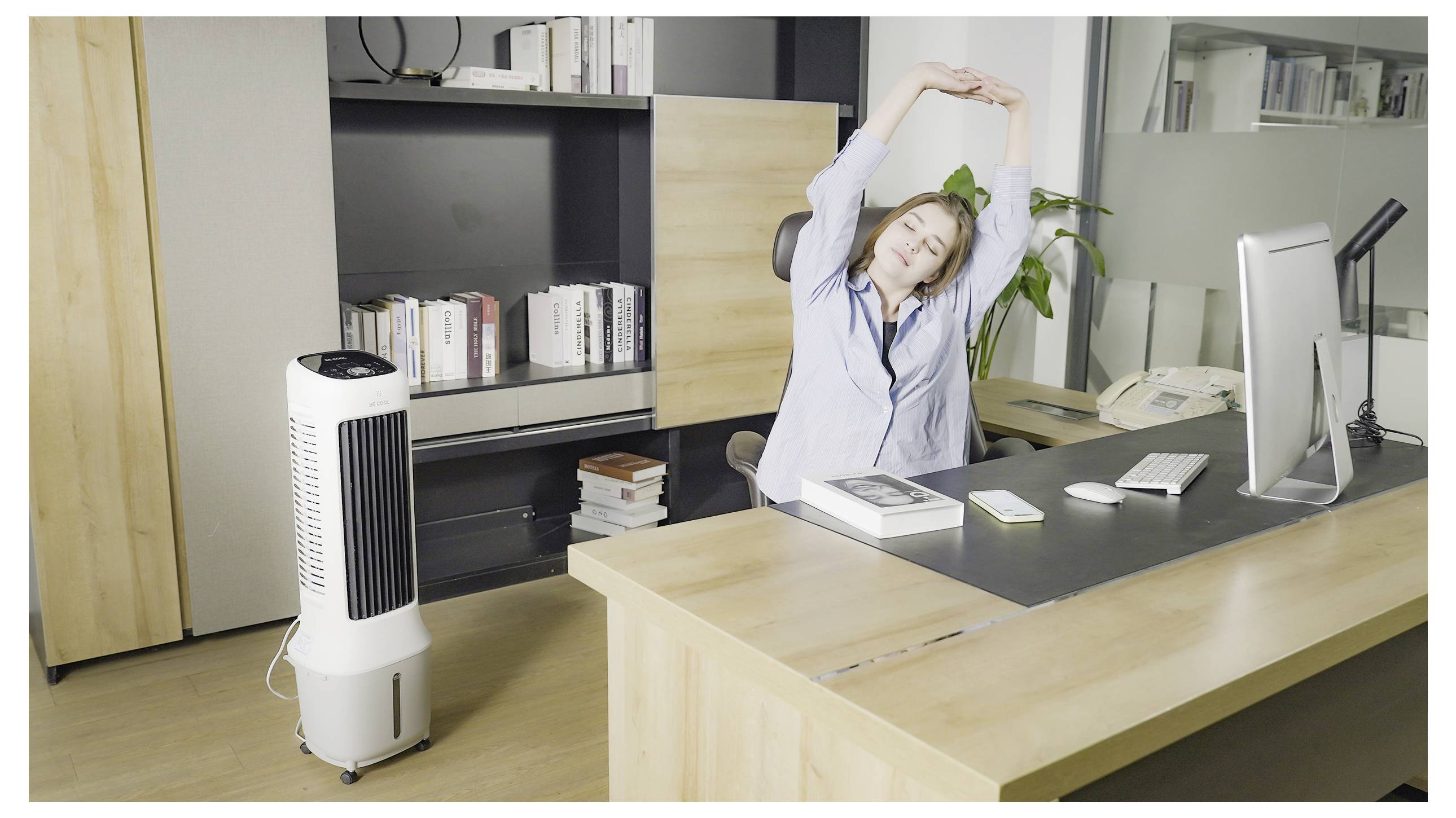 Eine Person, die an einem Schreibtisch im Büro sitzt, dehnt sich mit den Armen über dem Kopf aus, neben einem Ventilator, was auf eine Pause oder einen Entspannungsmoment hindeutet.