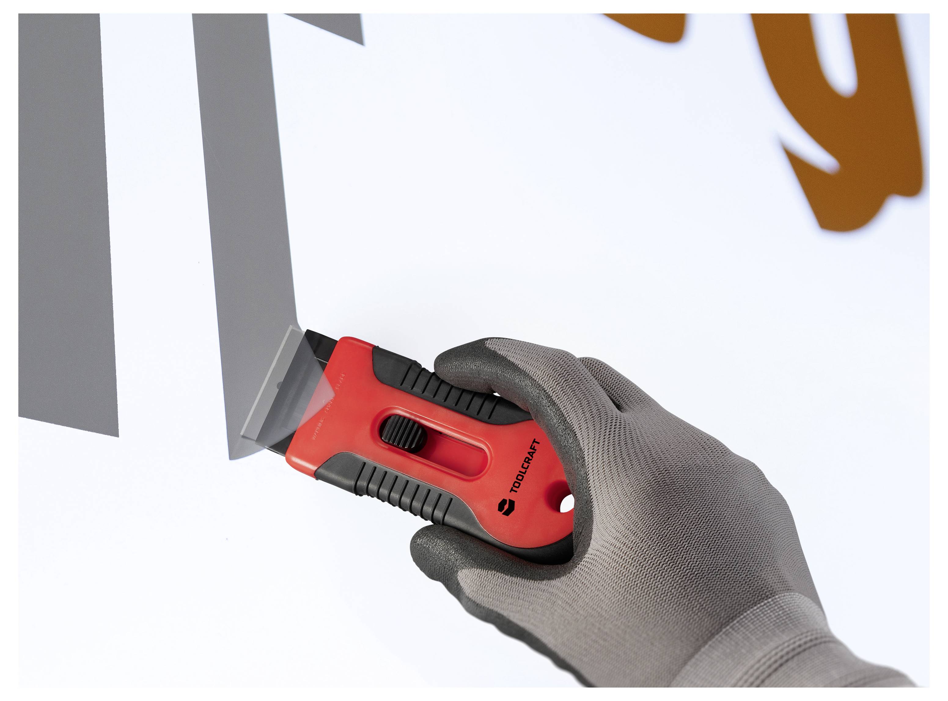 Eine behandschuhte Hand verwendet einen roten Sicherheitsmesser, um einen grauen Klebstoffbuchstaben von einer weißen Oberfläche abzulösen.