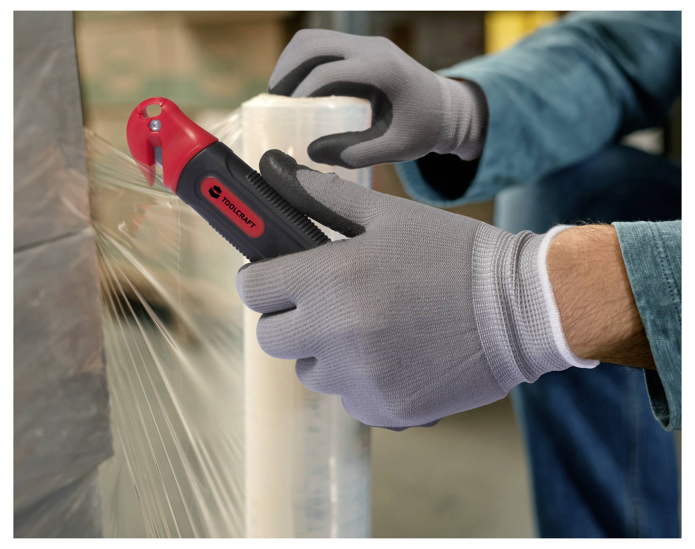 Eine Person, die graue Handschuhe trägt, verwendet einen rot-schwarzen Sicherheitsschneider, um eine Rolle durchsichtige Plastikfolie in einer Industrieumgebung zu schneiden.
