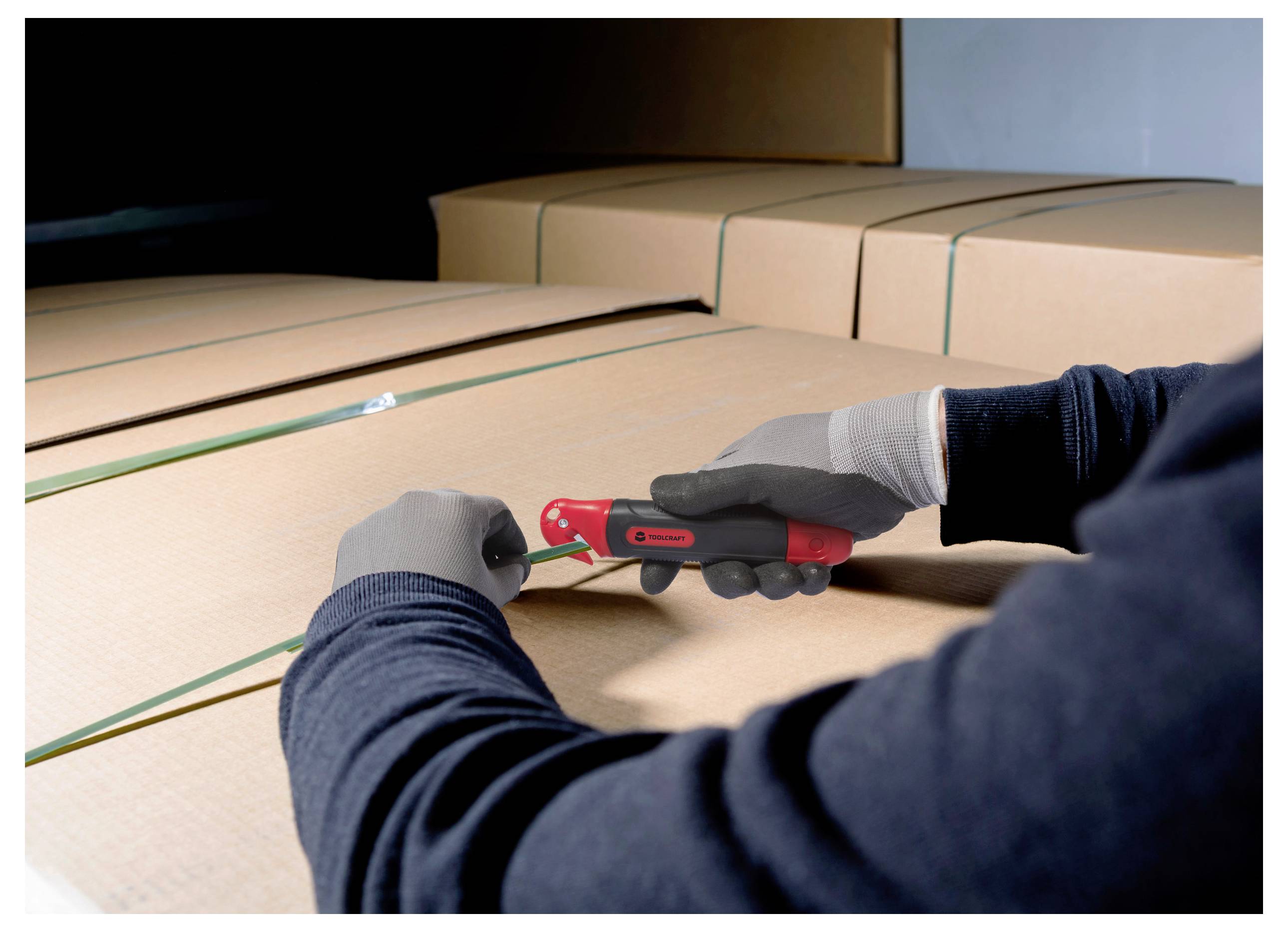 Eine Person trägt Handschuhe und benutzt einen roten Sicherheitsschneider, um die Kunststoffumreifung auf einem Stapel großer Kartonboxen in einem Lagerhaus zu durchschneiden.