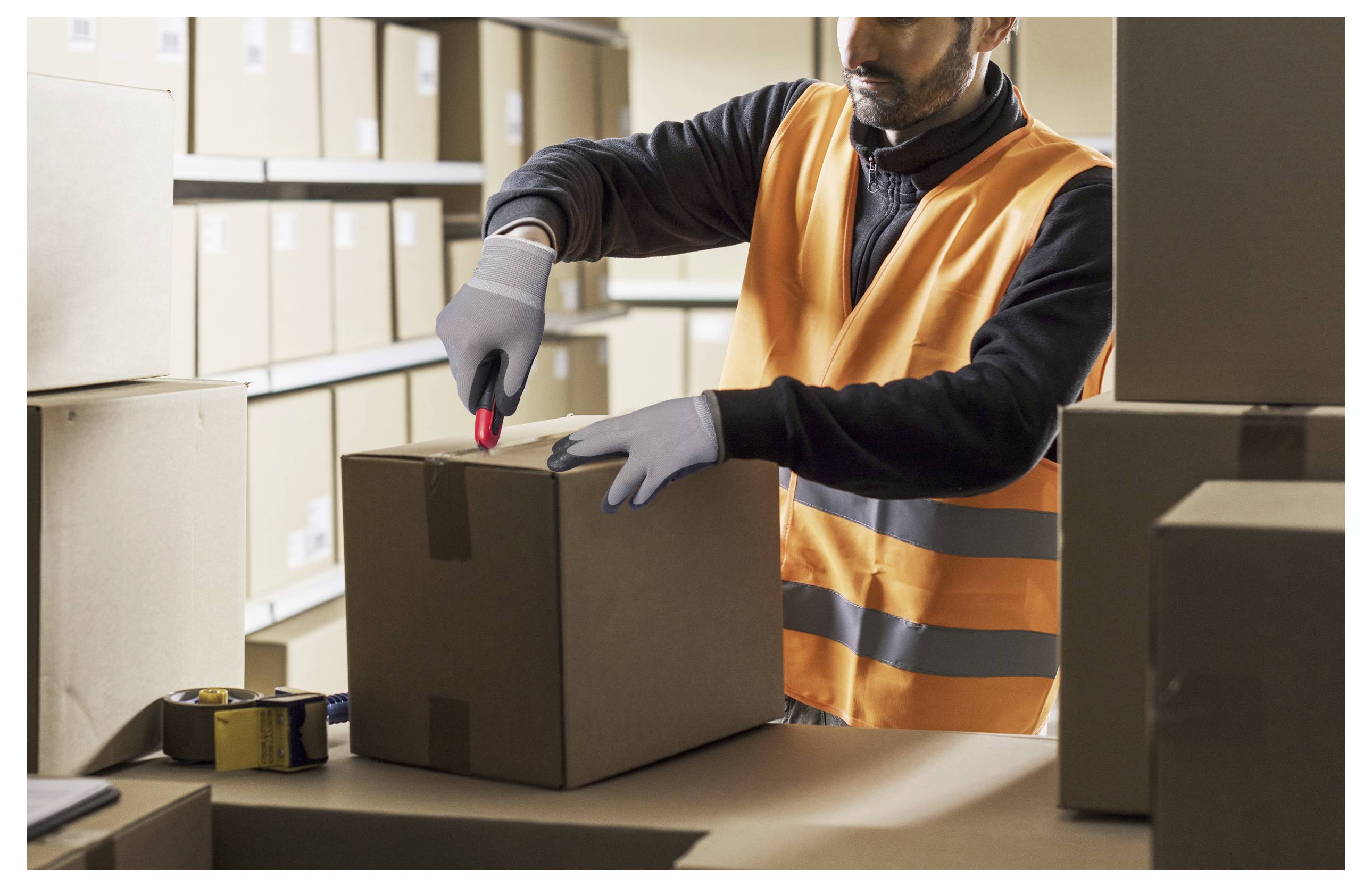 Ein Arbeiter in einer orangefarbenen Weste versiegelt eine Kartonschachtel mit Klebeband in einem Lagerbereich, der mit gestapelten Kartons gefüllt ist.