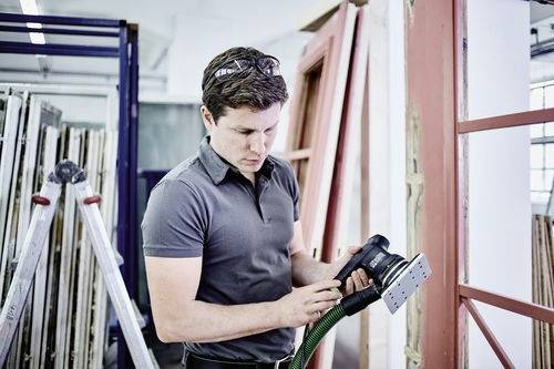 Eine Person in einer Werkstatt, die einen Elektro-Schleifer hält, scheint Holzrahmen vorzubereiten oder zu reparieren. Im Hintergrund befinden sich Leitern und Rahmen.