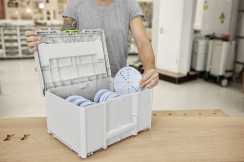 Eine Person legt eine Schleifscheibe in eine Aufbewahrungsbox, die bereits mit ähnlichen Scheiben gefüllt ist und auf einer Holzwerkbank in einer Werkstattumgebung steht.