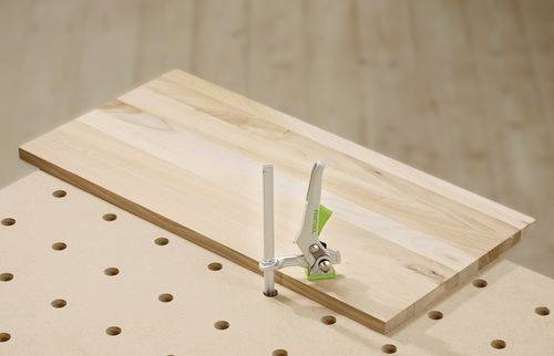 Ein Holzbrett, das mit einer Metallklemme auf einer gelochten Werkbank befestigt ist und für Holzarbeiten oder Schreineraufgaben verwendet wird.