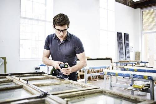 Eine Person mit Sicherheitsbrille verwendet ein Elektrowerkzeug, um einen Holzfensterrahmen in einer Werkstattumgebung zu restaurieren oder zu bearbeiten.