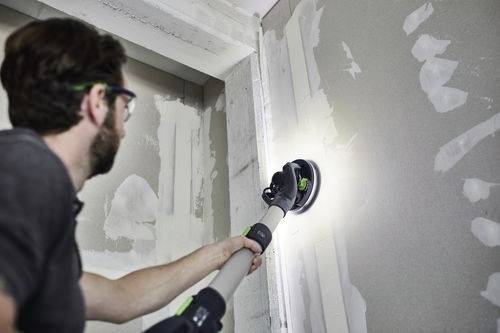 Eine Person verwendet ein Elektrowerkzeug, um Trockenbauwände in einem Raum im Bau zu schleifen und konzentriert sich dabei auf eine Naht zwischen zwei Platten.