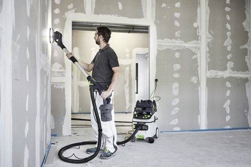 Eine Person schleift eine Wand mit einem langstieligen elektrischen Schleifgerät, das an einen Staubsauger angeschlossen ist, in einem teilweise fertiggestellten Raum mit Trockenbau.