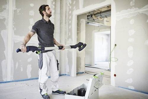 Ein Arbeiter, der in einem teilweise fertiggestellten Raum steht und ein Schleifwerkzeug hält, mit einer Werkzeugkiste auf dem Boden und Wänden, an denen unverkleidete Trockenbauplatten sichtbar sind.