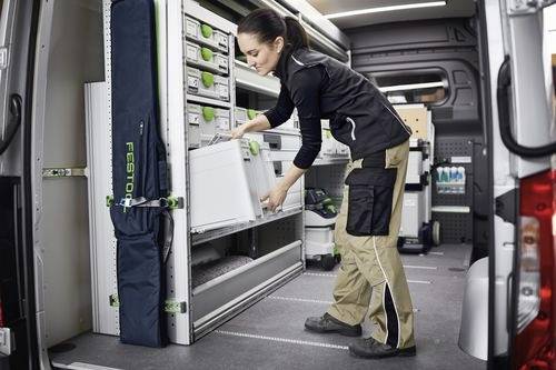 Eine Person richtet ein Lagersystem im Inneren eines Arbeitsfahrzeugs ein und zieht eine beschriftete Schublade heraus. Der Transporter ist mit Regalen und Werkzeugen ausgestattet.