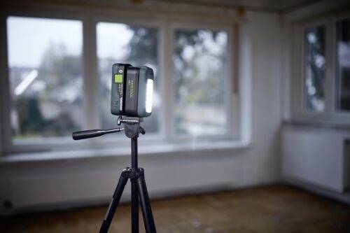Ein dreibeinig montierter Fotoapparat mit einem hellen Licht steht in einem leeren Raum mit großen Fenstern, was auf einen Fokus auf Fotografie oder Filmaufnahmen hindeutet.
