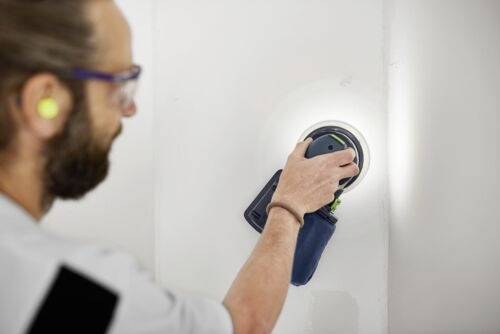 Eine Person benutzt einen Kreisschleifer an einer weißen Wand, trägt Schutzbrille und Gehörschutzstöpsel und konzentriert sich darauf, die Oberfläche zu glätten.