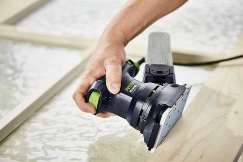 Eine Person benutzt einen Exzenterschleifer, um ein Holzstück auf einer Werkbank zu glätten. Das Werkzeug ist schwarz und grün mit einem Staubsammelaufsatz.
