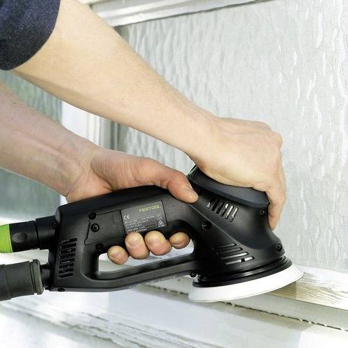 Eine Person verwendet eine elektrische Schleifmaschine an einem Holzfensterrahmen und glättet die Oberfläche. Die Schleifmaschine ist schwarz und hat ein sichtbares Netzkabel.