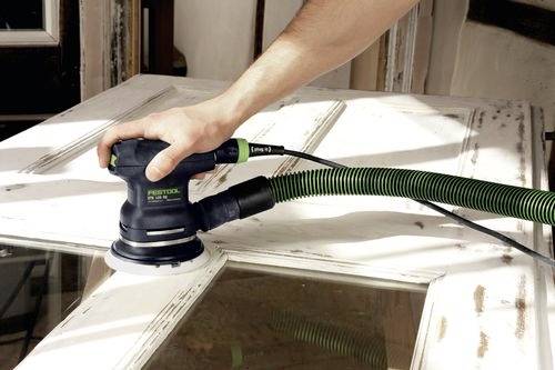 Eine Person verwendet eine elektrische Schleifmaschine an einer weißen Holztür und glättet die Oberfläche. Die Szene deutet auf Heimwerken oder Holzbearbeitung hin.