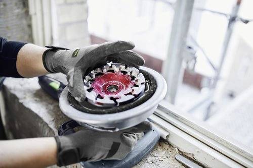 Eine Person mit Schutzhandschuhen hält und untersucht ein kreisförmiges Schleifwerkzeug in der Nähe eines Fensters, was auf eine Bau- oder Renovierungsumgebung hindeutet.