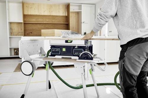 Eine Person, die an einer Kreissäge arbeitet und eine Holzplatte in einem hell erleuchteten Raum mit Regalen zersägt. Sicherheitsausrüstung ist auf dem Tisch sichtbar.