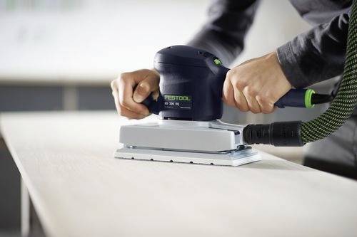 Eine Person benutzt einen elektrischen Schleifer auf einer Holzoberfläche und glättet sie. Der Schleifer ist schwarz mit grünen Akzenten und an einen Schlauch angeschlossen.