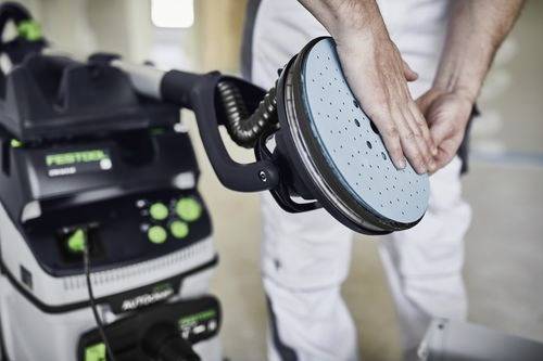 Eine Person justiert einen blauen Schleifpad an einer Schleifmaschine und bereitet sich auf eine Holzbearbeitungsaufgabe vor. Verschiedene Bedienelemente und Einstellungen sind sichtbar.