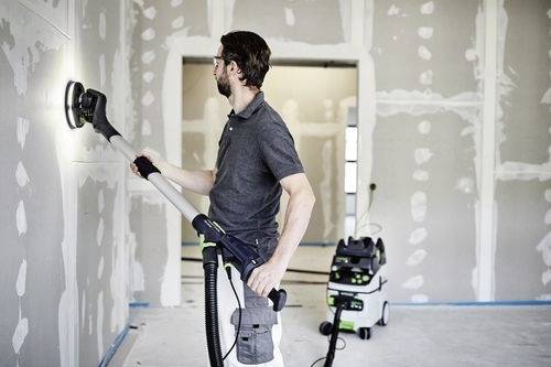 Eine Person verwendet einen Trockenbauschleifer an einer Wand in einem Raum, der sich im Bau befindet. Ein Vakuumsystem ist mit dem Schleifer zur Staubkontrolle verbunden.