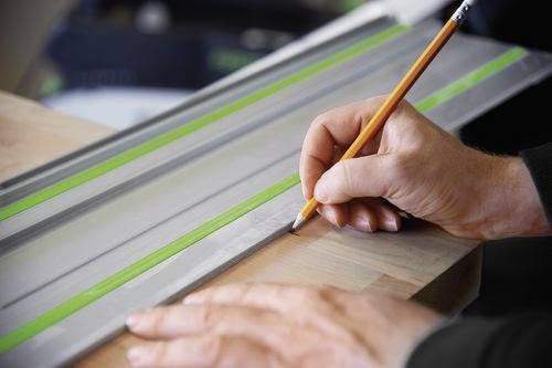 Eine Person verwendet einen Bleistift, um ein Holzbrett mit einem grün gestreiften Metalllineal zu markieren und zeigt dabei präzise Messung und Planung.