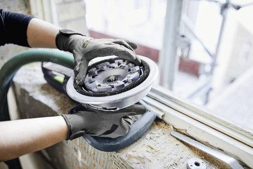 Arbeiter mit Handschuhen verwendet einen elektrischen Schleifer an einem Fensterrahmen und glättet die Oberfläche in der Nähe einer Baustelle.