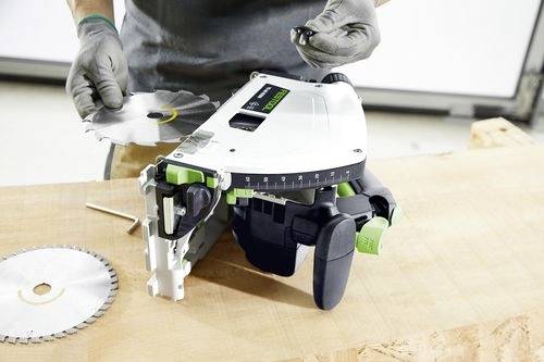 Eine Person montiert eine Kreissäge auf einer Holzwerkbank, hält eine Sägeblatt und ein kleines Anbauteil, wobei Werkzeuge in der Nähe sichtbar sind.