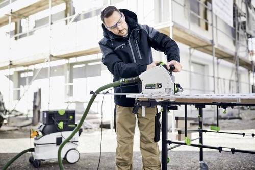 Mann, der eine Festool-Säge im Freien auf einer Baustelle verwendet und Schutzausrüstung trägt. Gerüst und Gebäudefassade im Hintergrund.