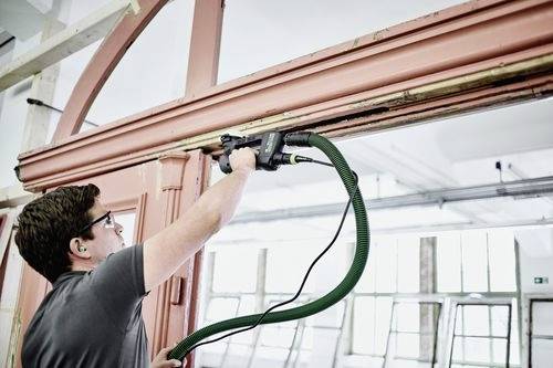 Eine Person verwendet ein Elektrowerkzeug, um einen Türrahmen zu schleifen, wobei ein grüner Schlauch zur Staubabsaugung angeschlossen ist, in einem hell erleuchteten Raum mit großen Fenstern.