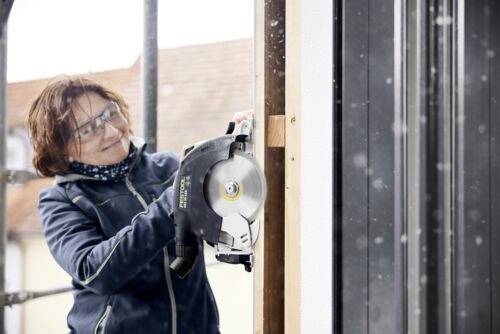 Eine Person mit Schutzbrille verwendet eine Kreissäge, um ein Holzbrett im Freien zu sägen.