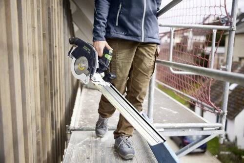 Eine Person steht auf Gerüstbohlen und hält eine Führungskreissäge, trägt Sicherheitsausrüstung. Die Umgebung ist eine Baustelle im Freien in erhöhter Position.