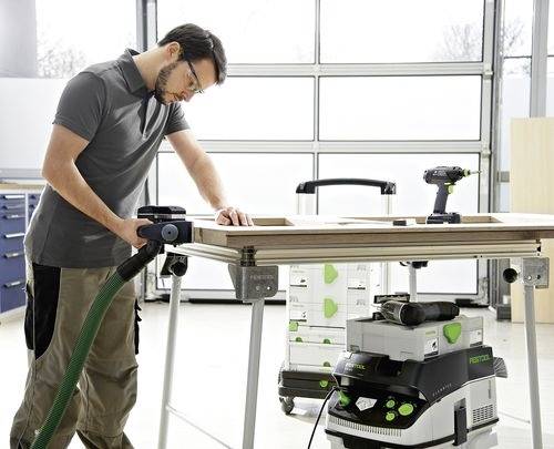 Eine Person mit Schutzbrille schleift ein Brett mit einem Elektrowerkzeug in einer Werkstatt. Ein Staubabsauger ist angeschlossen und sorgt für einen sauberen Arbeitsbereich.