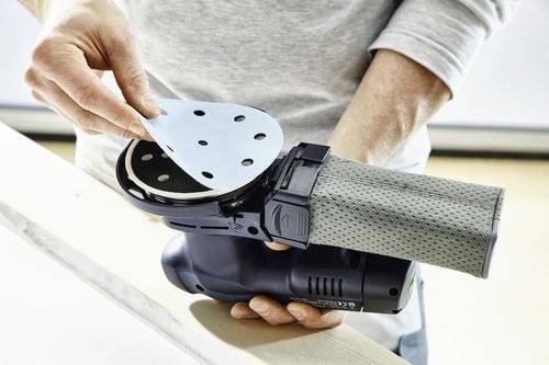 Eine Person bereitet eine tragbare Elektroschleifmaschine vor, indem sie eine scheibenförmige Schleifunterlage anbringt. Die Schleifmaschine verfügt über einen Staubbeutel zur Staubsammlung.