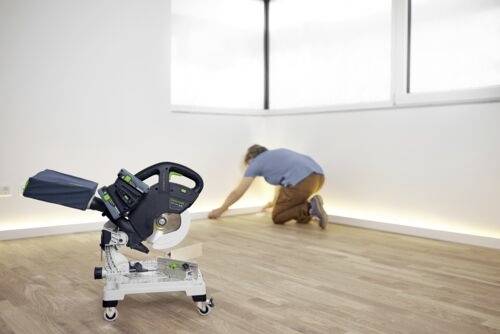 Eine Person bringt Fußleisten entlang einer weißen Wand in einem Raum mit Holzfußboden an. Eine Kapp- und Gehrungssäge ist auf dem Boden positioniert.