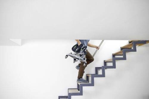 Eine Person trägt einen auf den Kopf gestellten Bürostuhl und steigt vorsichtig eine schmale, moderne Holztreppe mit einer weißen Wand im Hintergrund hinab.